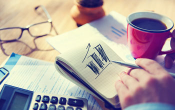 A hand drawing a graph with 3 vertical lines on it. The pad is on a desk with a calculator , coffee and glasses