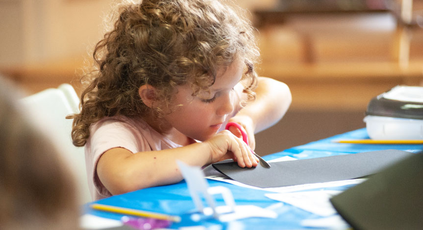 A girl sitting at a desk using scissors to cut a black piece of black construction paper