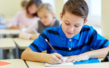 A boy sitting at his desk writing in his note book using a pencil